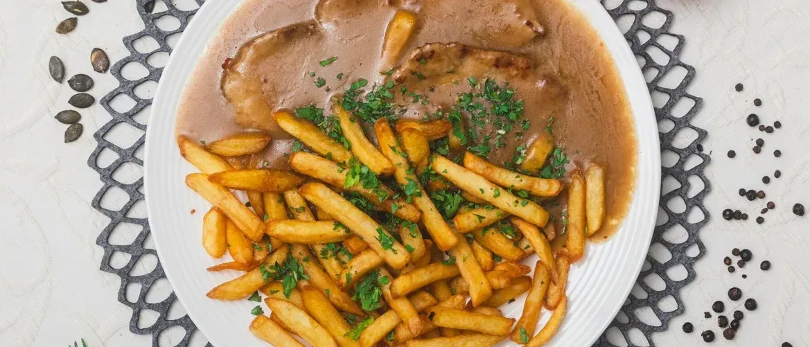 Ein Teller mit Pommes Frites, übergossen mit Soße und Petersilie, begleitet von zwei Fleischstücken.