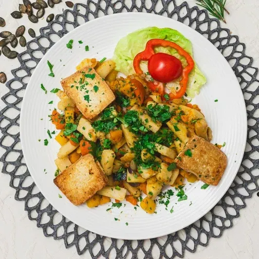 Ein weißer Teller mit einer Mahlzeit aus gerösteten Gemüse, garniert mit Petersilie, einer Scheibe roter Paprika und einigen Stücken gebratenem Brot.