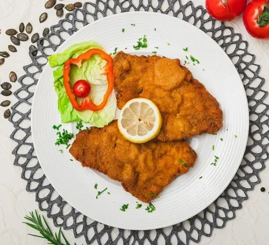 Ein Teller mit panierten Hühnchenschnitzeln mit einer Zitronenscheibe, serviert mit einem Salat aus Salat, Paprika und Kräutern, auf einer gemusterten Tischdecke.