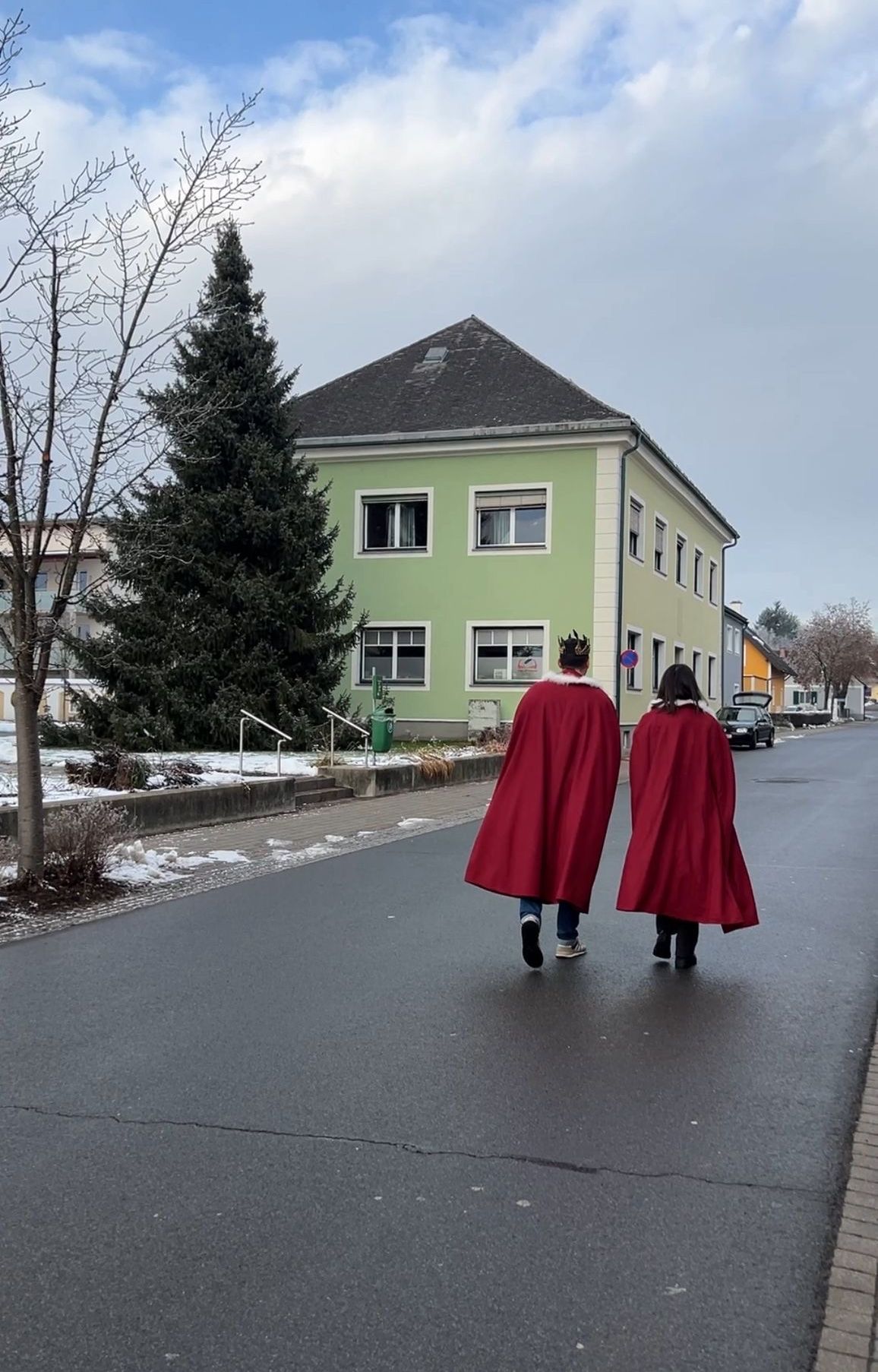 Zwei Personen in roten Umhängen gehen vor einem grünen Gebäude mit einem Baum auf einer verschneiten Straße.