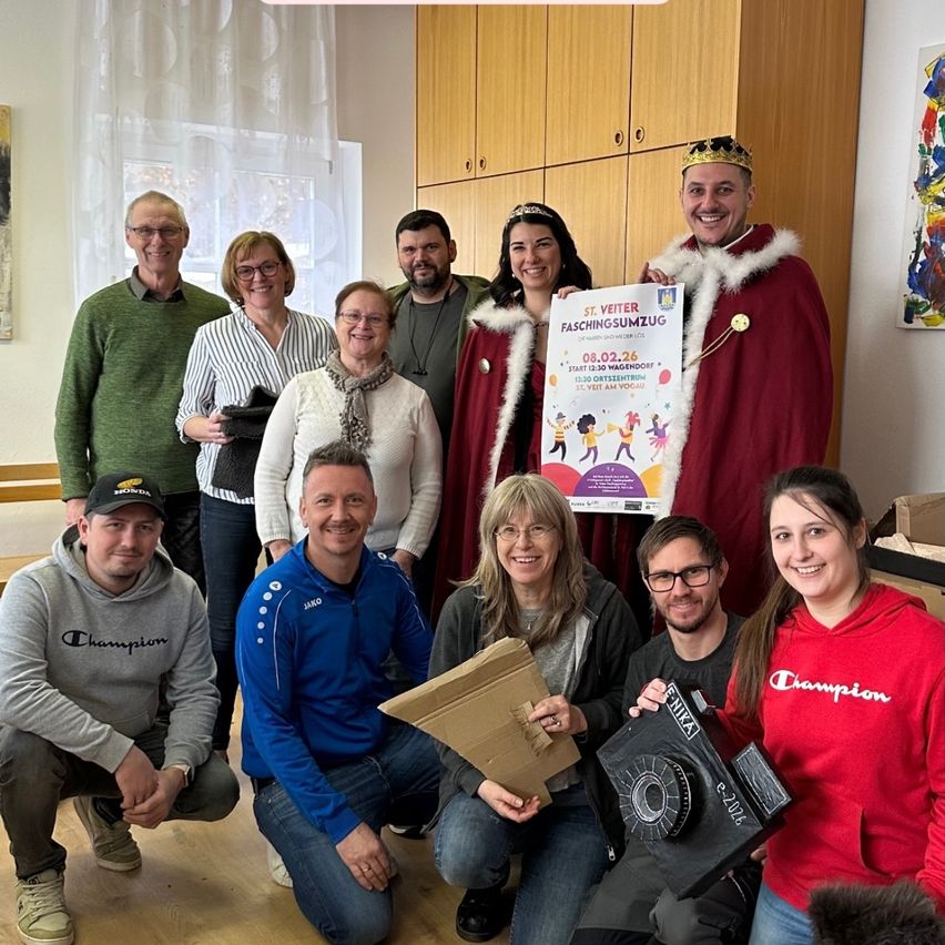 Eine Gruppe von Menschen posiert für ein Foto in einem Raum, einige halten Papierboxen. Eine Frau mit Krone und ein Mann in rotem Umhang halten ein Plakat. Sie alle lächeln.