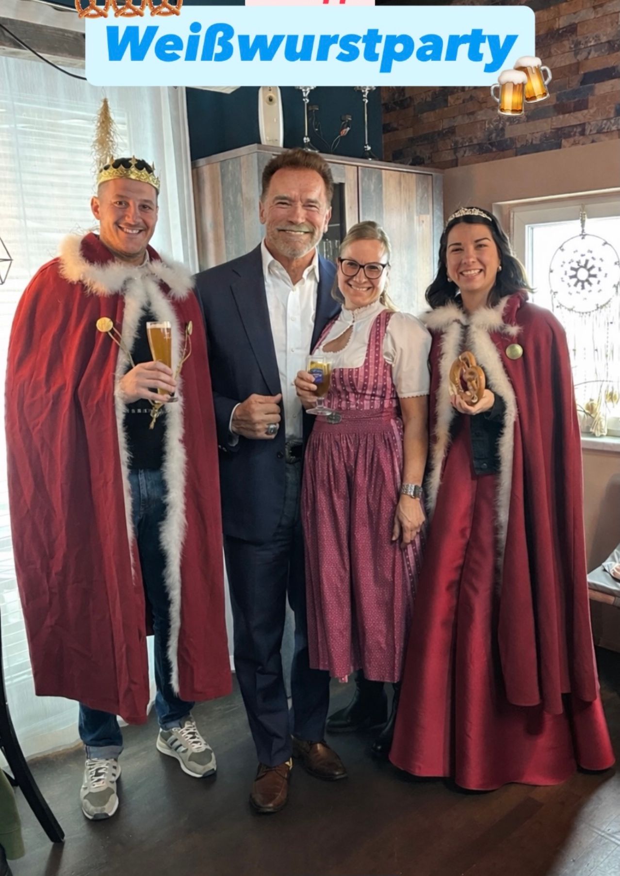 Vier Personen in festlicher Kleidung, die Getränke in der Hand halten und für ein Foto lächeln. Der Mann in der Mitte trägt eine Krone und einen Anzug. Die beiden Frauen in roten Umhängen halten Brezen.