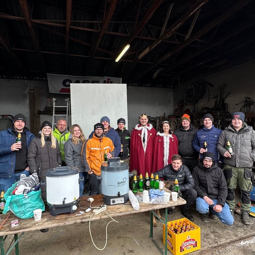 Eine Gruppe von Menschen versammelt sich in einer Garage mit verschiedenen Gegenständen auf einem Tisch, einschließlich Flaschen, Tassen und einem Hund. Einige tragen Winterkleidung, und einer ist in einer Weihnachtsmann-Kostümierung.