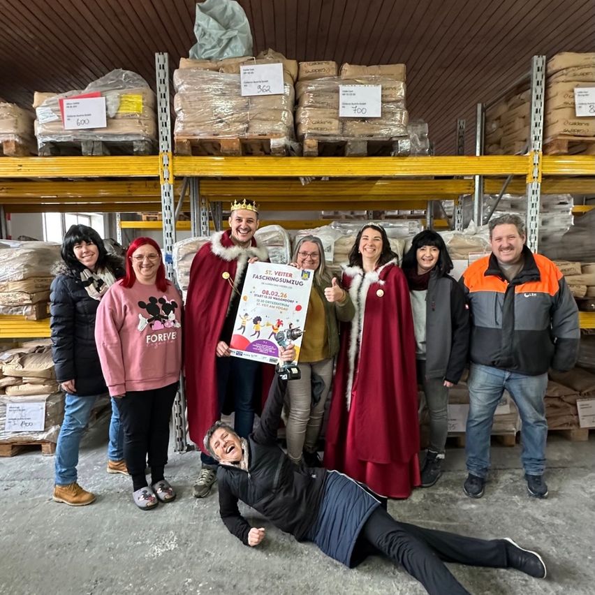 Eine Gruppe von Menschen steht in einem Lagerhaus, einige in Kostümen. Ein Mann hält ein Plakat und ein Mikrofon, während andere lächeln und posieren. Sie sind von gestapelten Kisten umgeben.