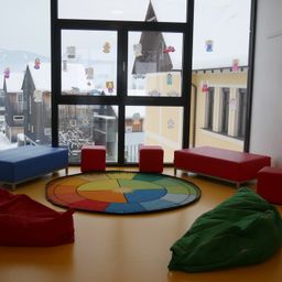 Ein gemütlicher Raum mit einem bunten Teppich, Sitzsäcken und Hockern, mit einem Fenster, das eine verschneite Landschaft draußen zeigt.
