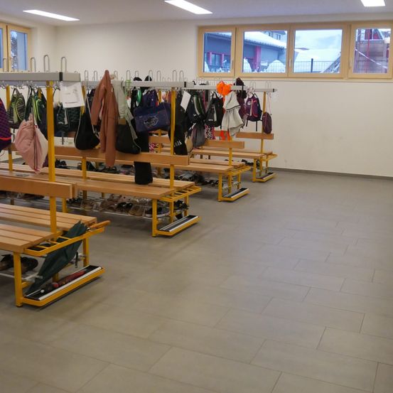 Ein leerer Raum mit Bänken und Kleiderhaken für Mäntel und Taschen, beleuchtet durch Deckenleuchten und natürliches Licht aus Fenstern.