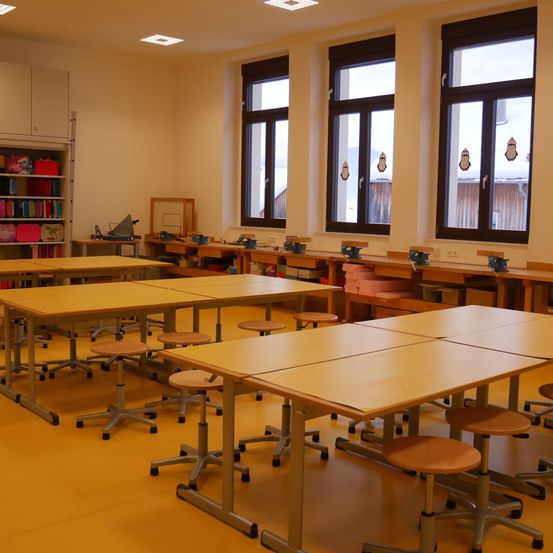 Ein leerer Klassenraum mit hölzernen Tischen und Stühlen in Reihen, großen Fenstern mit Pinguinaufklebern und einem Bücherregal voller bunter Bücher.