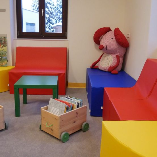 Ein Spielzimmer mit bunten Stühlen und einem Holzwagen voller Bücher. Ein Plüschtier sitzt auf einem blauen Stuhl.