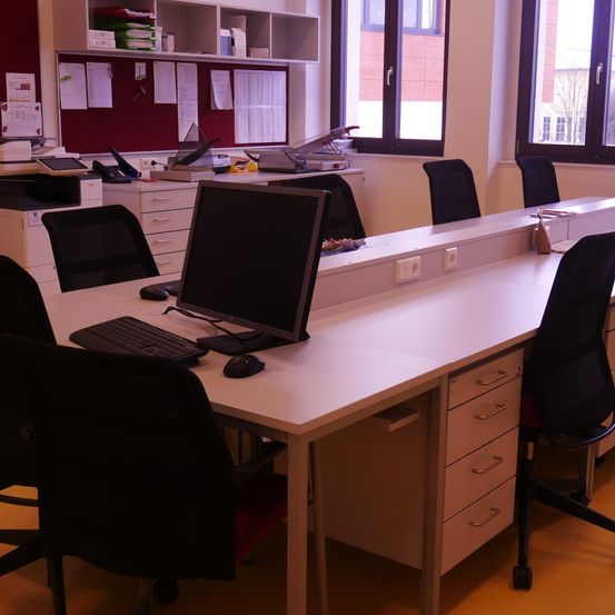 Ein Büro mit mehreren Schreibtischen, jeder mit einem Computermonitor, einer Tastatur und einer Maus ausgestattet. Der Raum hat große Fenster und eine Anschlagtafel mit angehefteten Papieren.