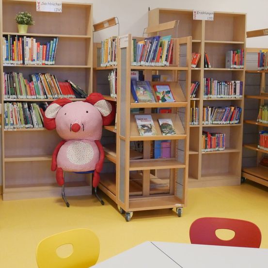 Eine Bibliothek mit Holzregalen voller Bücher und einem Plüschtier, das auf einem Stuhl sitzt. Der Boden ist gelb, und es gibt Stühle und einen Tisch im Raum.