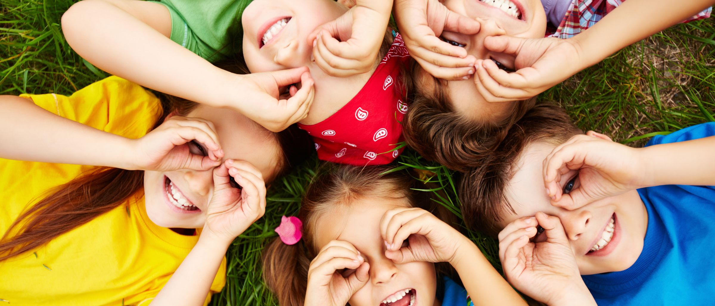 Eine Gruppe von Kindern liegt auf dem Gras und bildet mit den Händen über den Augen einen Kreis. Sie lächeln und haben Spaß.