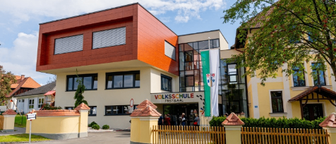Das Volksschule Fürstsaal-Gebäude mit seiner modernen Architektur ist von einem gut gepflegten Garten und einem Holzzaun umgeben. Das Gebäude weist eine Mischung aus roten und gelben Außenfassaden auf, und es befinden sich mehrere Personen am Haupteingang.