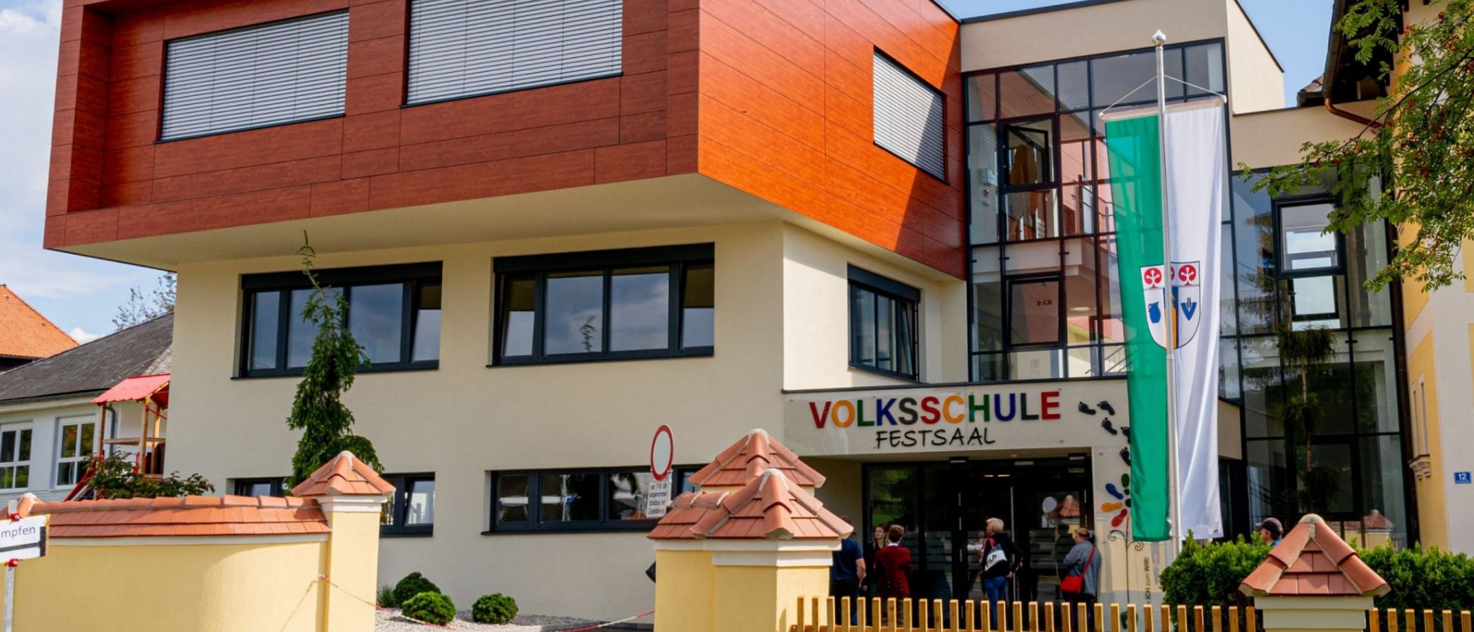 Ein modernes Gebäude mit roter Fassade und Glasfenstern, mit einer Flagge und einem Schild, das 'Volksschule Festsaal' besagt. Der Eingang ist von einem Holzzaun und einem Rasen umgeben.