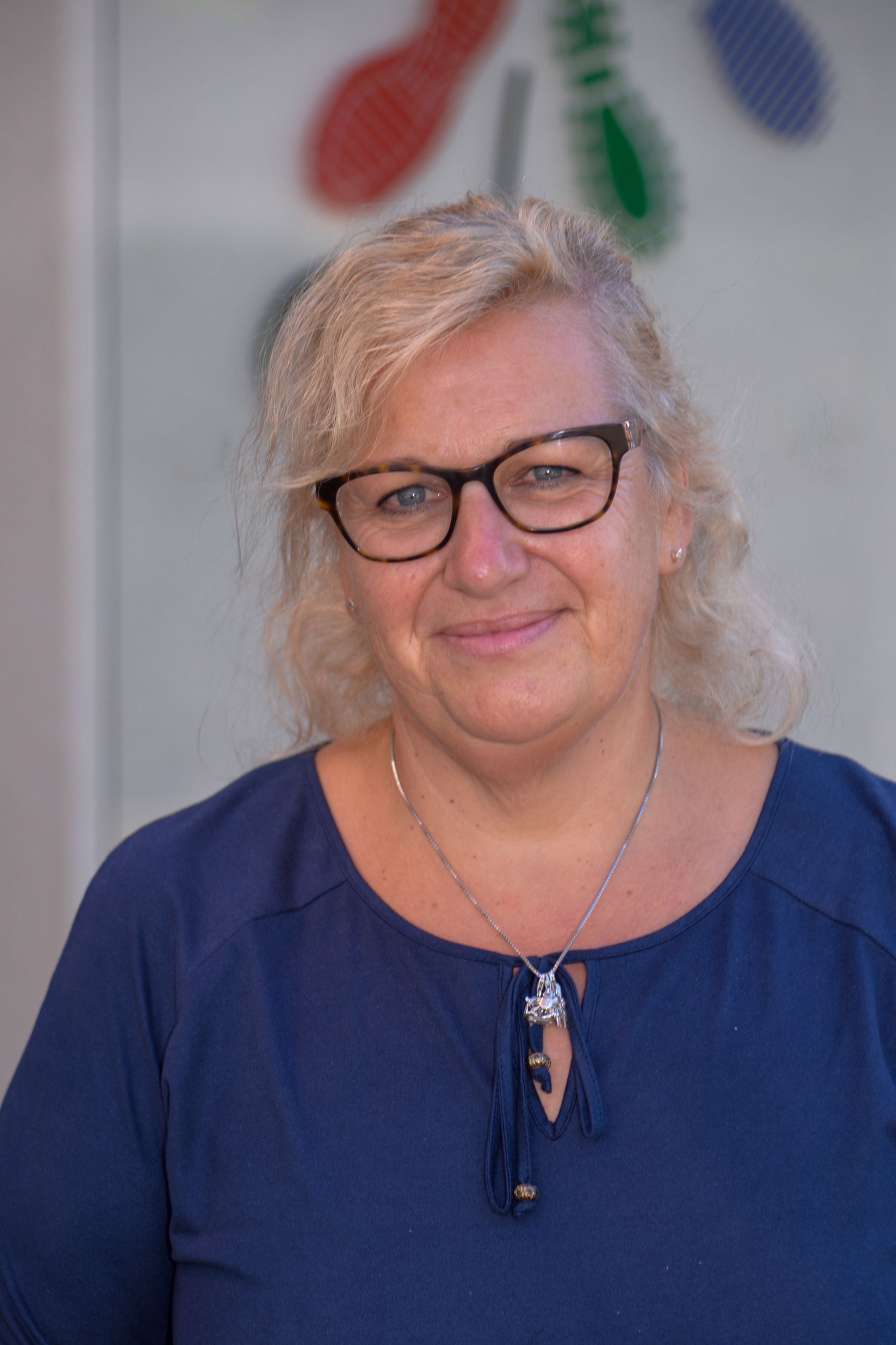 Eine Frau mit blonden Haaren, die eine Brille und ein blaues Hemd trägt, lächelt für die Kamera. Sie hat eine Halskette mit einem Anhänger.