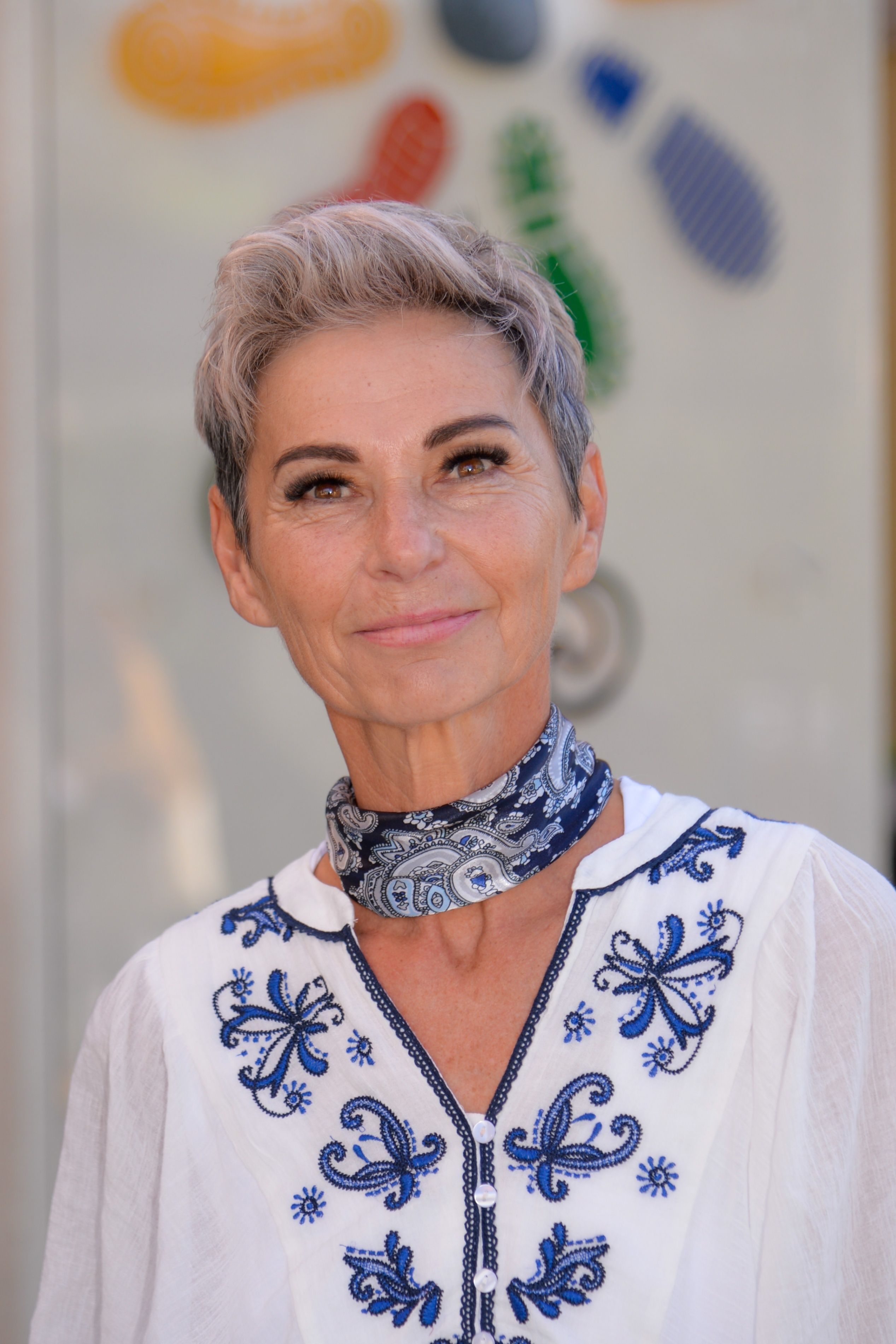Eine Frau mit kurzen grauen Haaren und einem blauen Paisley-Schal lächelt in die Kamera. Sie trägt ein weißes Oberteil mit blauer Stickerei.