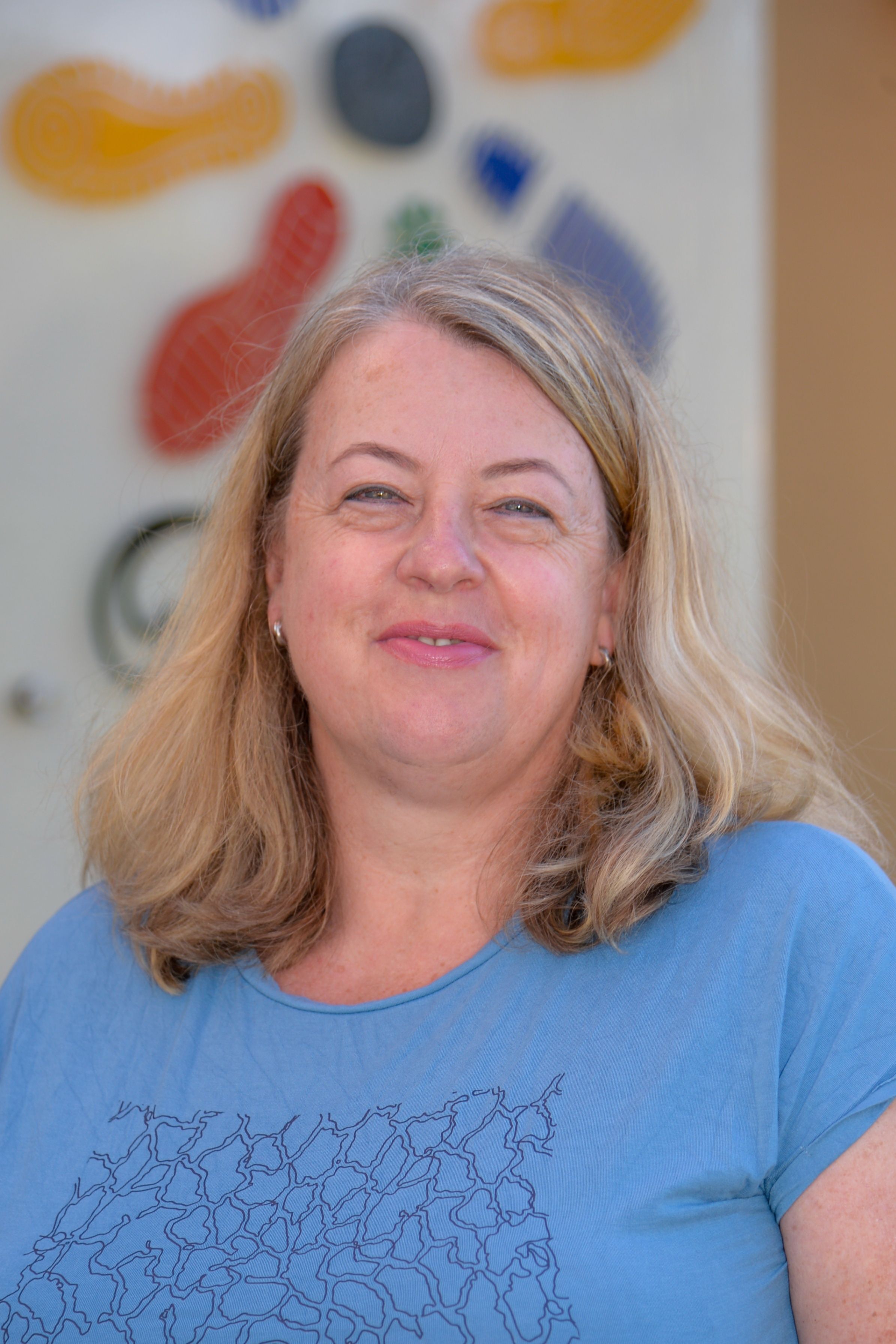 Eine Frau mit blonden Haaren und einem blauen T-Shirt lächelt in die Kamera. Sie hat einen Muttermal auf ihrer rechten Wange.