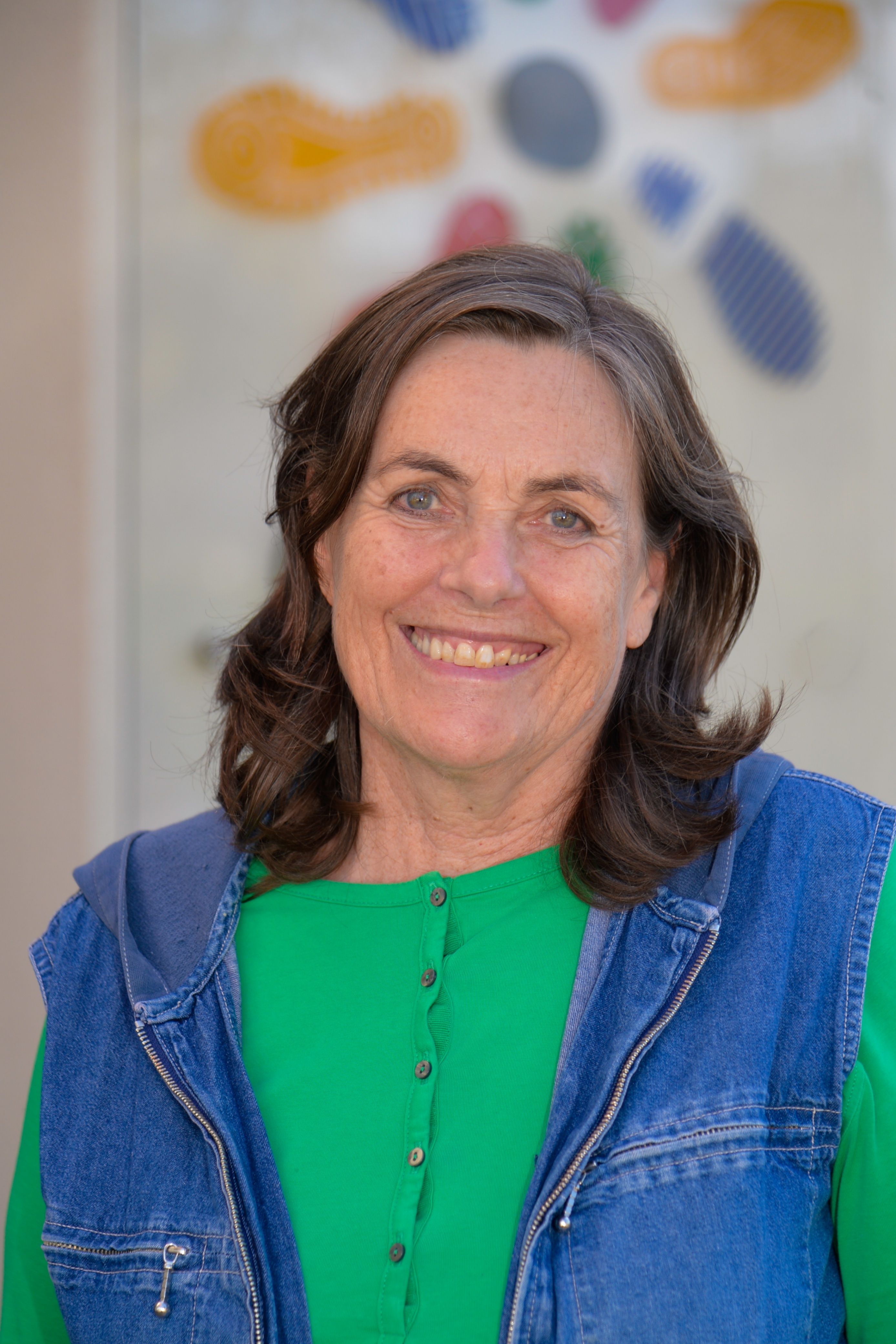 Eine Frau mit braunen Haaren, die ein grünes Shirt und eine blaue Jeansweste trägt, lächelt in die Kamera.