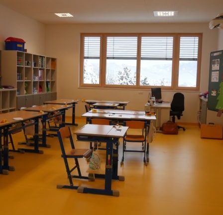 Ein leerer Klassenraum mit gelbem Boden, Holztischen und Stühlen. Es gibt Regale mit Büchern und Aufbewahrungsboxen, ein Fenster mit Jalousien und ein Projektor an der Decke.