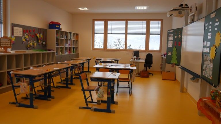 Ein leerer Klassenraum mit gelbem Boden, Holztischen und Stühlen. Es gibt Regale mit Büchern und Aufbewahrungsboxen, ein Fenster mit Jalousien und ein Projektor an der Decke.