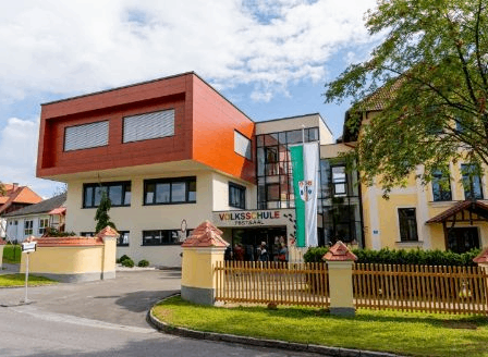 Das Volksschule Postsaal-Gebäude ist ein modernes architektonisches Bauwerk mit rotem, gelbem und weißem Äußeren. Es hat eine Flagge am Eingang. Bäume und Pflanzen umgeben das Gebäude.