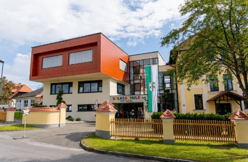 Das Volksschule Postsaal-Gebäude ist ein modernes architektonisches Bauwerk mit rotem, gelbem und weißem Äußeren. Es hat eine Flagge am Eingang. Bäume und Pflanzen umgeben das Gebäude.