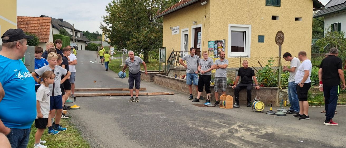 Eine Gruppe von Menschen, einschließlich Kindern, versammelt sich auf einer Straße. Ein erwachsener Mann wirft einen Ball in Richtung eines Ziels. Andere Leute stehen und beobachten. Ein gelbes Haus mit Balkon und Garten ist sichtbar.