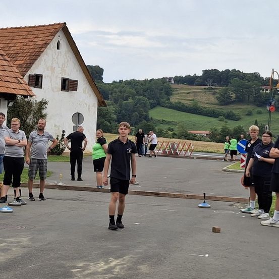Menschen spielen ein Spiel im Freien. Ein Junge bereitet sich darauf vor, einen Scheiben zu werfen, während andere zusehen. Gebäude und Bäume sind im Hintergrund.