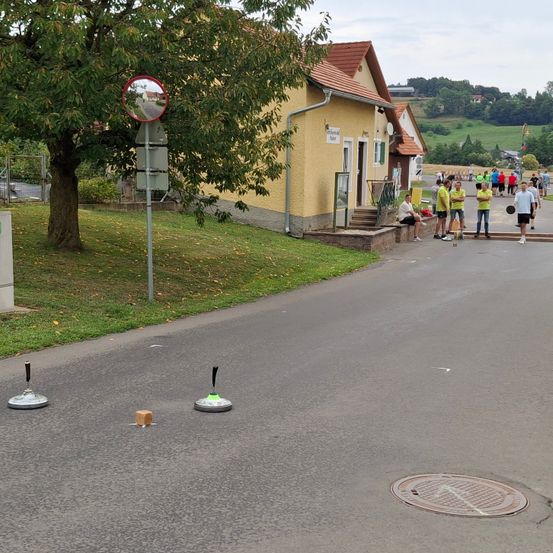Eine Außenszene mit einer Straße, auf der Kegel, ein runder Spiegel an einem Pfahl, eine Gruppe von Menschen und ein Gebäude mit Treppen im Hintergrund zu sehen sind.