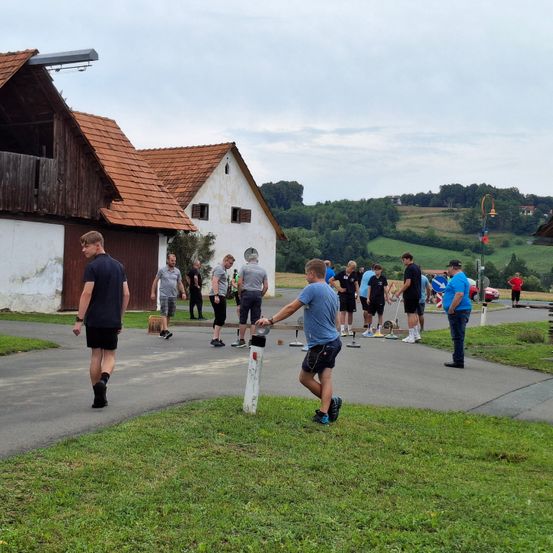 Eine Gruppe von Menschen ist auf einem Hof versammelt. Ein Mann in einem blauen Shirt wirft einen Ball. Hinter ihm stehen mehrere Leute und schauen zu. Die Szene spielt vor dem Hintergrund eines rustikalen Gebäudes.
