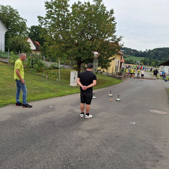 Zwei Männer stehen auf einer Straße, einer trägt ein gelbes Shirt und der andere ist in Schwarz gekleidet. Sie befinden sich in der Nähe eines Rasens mit Bäumen. In der Ferne gehen Menschen auf der Straße.