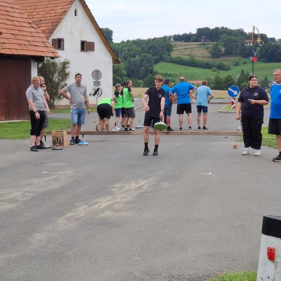Eine Gruppe von Menschen spielt ein Spiel auf einer Betonfläche. Ein Mann wirft einen Frisbee. Einige Leute stehen, während andere sich bücken.