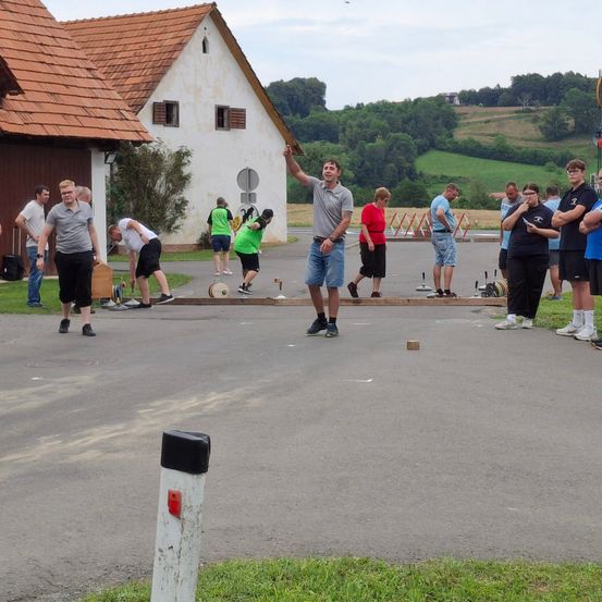 Eine Gruppe von Menschen nimmt draußen an einem Spiel teil, wobei einige Turnschuhe tragen und andere im Kreis stehen. Ein Mann wirft ein Objekt. Bäume und ein Haus sind im Hintergrund.