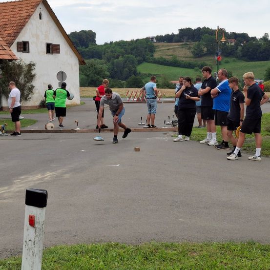Eine Gruppe von Menschen versammelt sich vor einem Gebäude und ist mit einer kleinen Aktivität am Boden beschäftigt. Ein Mann in einem grauen Shirt bereitet sich darauf vor, es zu werfen. Zuschauer beobachten, einige tragen grüne Shirts, während andere verschiedene Farben tragen. Ein Grasbereich und ein Gebäude mit einem roten Dach sind im Hintergrund.