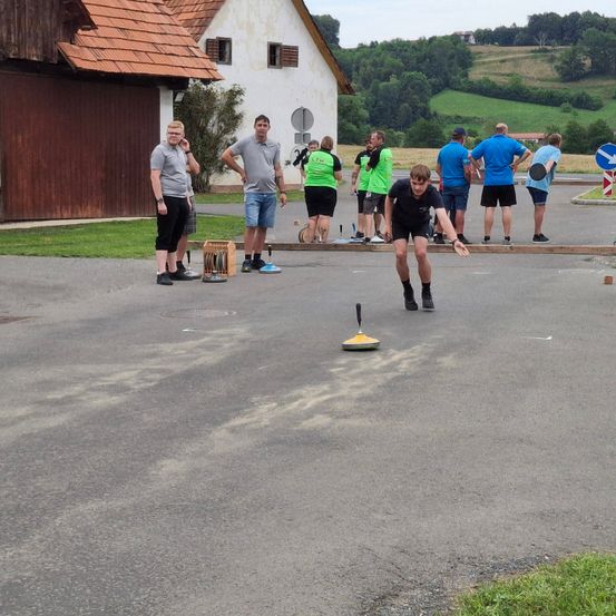 Eine Gruppe von Menschen spielt draußen an einem sonnigen Tag ein Spiel. Ein Junge in Schwarz wirft ein gelbes Objekt.