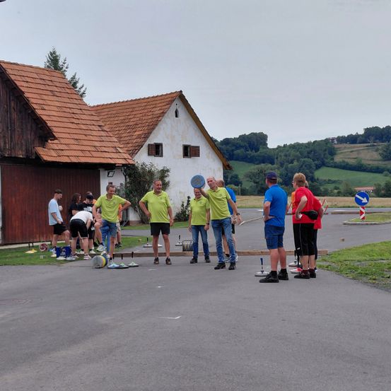 Eine Gruppe von Menschen in neongrünen Shirts steht in einer Reihe auf der Straße, möglicherweise in einem Spiel engagiert. Sie halten einen Frisbee und andere beugen sich in ihrer Nähe. Dahinter befindet sich ein Gebäude mit einem braunen Dach und einer weißen Wand.