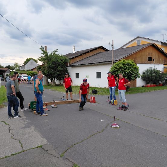 Mehrere Männer in roten Trikots spielen eine Partie Boccia auf einer offenen Fläche. Dahinter befinden sich Gebäude, ein Baum und ein Rasen.