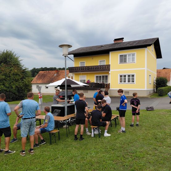 Eine Gruppe von Personen versammelt sich auf einem Rasen vor einem gelben Haus, mit Tischen und einem Schirm unter einem bewölkten Himmel. Einige sitzen, andere stehen.