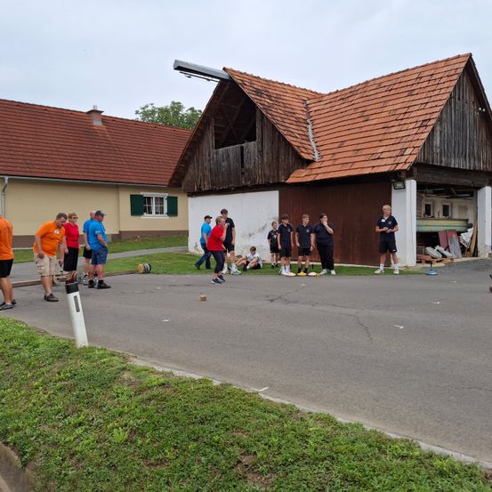 Eine Gruppe von Menschen steht vor einem hölzernen Gebäude mit einem braunen Dach, einige spielen. In der Nähe sitzt eine Person auf dem Rasen.