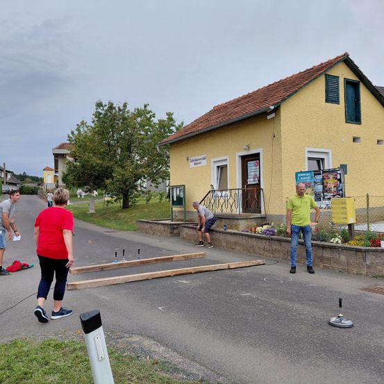 Eine Gruppe von Menschen arbeitet an einer Straße vor einem gelben Haus. Ein Mann beugt sich, während ein anderer neben einem Briefkasten steht. Eine Frau in einem roten T-Shirt und Turnschuhen schaut zu.