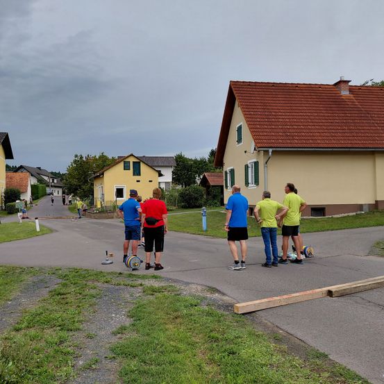 Eine Gruppe von Menschen in passenden blauen und gelben Shirts steht auf einem Betonweg, einige stehen in der Nähe einer Holzplanke. Mehrere Häuser mit braunen Dächern sind im Hintergrund zu sehen.
