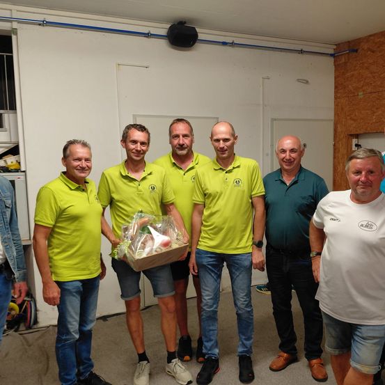 Fünf Männer stehen in einem Raum, lächeln und posieren für ein Foto. Sie tragen grüne und weiße Poloshirts, außer einem in blau. Ein Mann hält eine durchsichtige Box mit Essen. Der Raum hat weiße Wände und eine Holzwand.