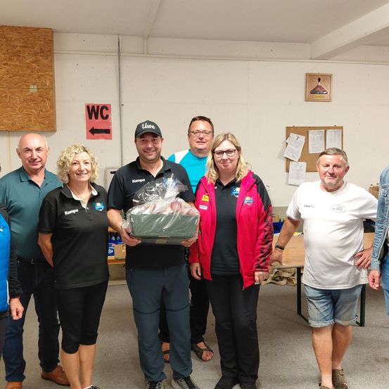 Eine Gruppe von Menschen steht lächelnd in einem Raum. Ein Mann mit einer Kappe hält ein verpacktes Geschenk. Hinter ihnen befindet sich ein 'WC'-Schild an der Wand und ein Tisch mit angehefteten Papieren.