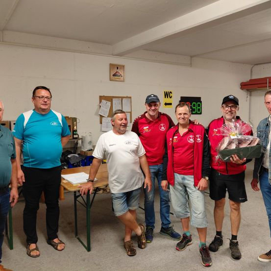 Fünf Männer stehen in einem Raum, lächeln und posieren für ein Foto. Sie tragen Sporttrikots und Freizeitkleidung. Ein Mann in der Mitte hält einen Geschenkkorb. Hinter ihnen steht ein Tisch mit einem Buch darauf.