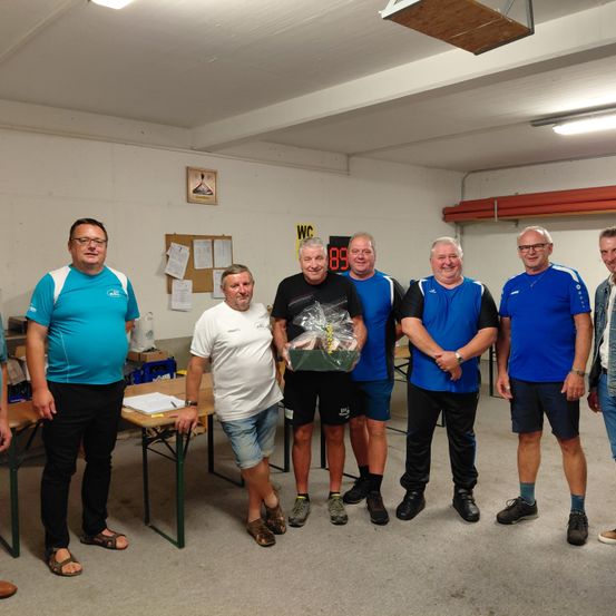 Eine Gruppe von Männern steht in einem Raum, einige lächeln. Ein Mann hält einen Geschenkkorb. Hinter ihnen sind Tische und Papiere zu sehen, sowie ein Bulletin-Board.