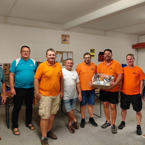 Sechs Männer stehen in einem Raum. Sie tragen orangefarbene Hemden und Shorts. Ein Mann hält einen Geschenkkorb. Sie alle lächeln. Im Hintergrund befindet sich ein Whiteboard und eine Digitaluhr.