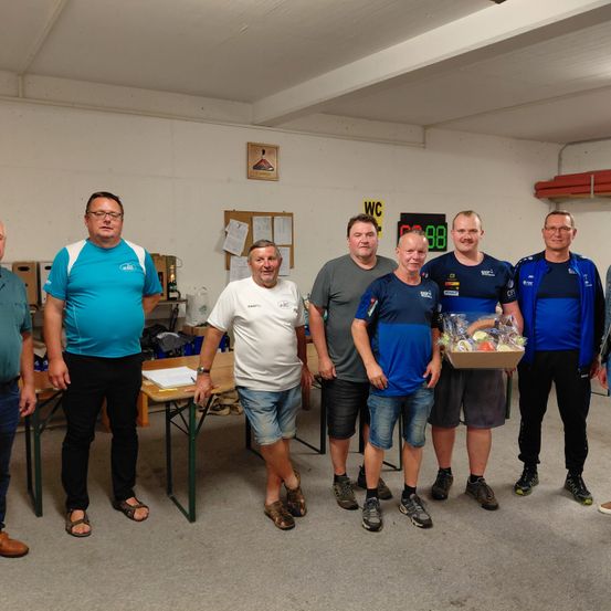 Eine Gruppe von Männern steht in einem Raum und lächelt für ein Foto. Sie tragen Freizeitkleidung. Einer hält eine Schachtel mit Gegenständen darin. Ein Tisch und Papiere sind hinter ihnen.