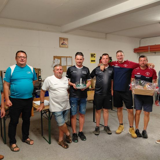 Sechs Männer stehen in einem Raum, drei halten eine Trophäe und zwei halten eine Geschenkbox. Sie tragen Freizeitkleidung und Turnschuhe. Hinter ihnen steht ein Tisch mit einer Flasche und ein Rahmen an der Wand.