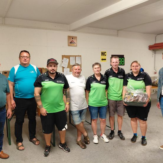 Sieben Personen in einem Raum, fünf von ihnen in grünen und schwarzen Shirts, lächeln für die Kamera. Sie halten eine Box mit verschiedenen Gegenständen darin.