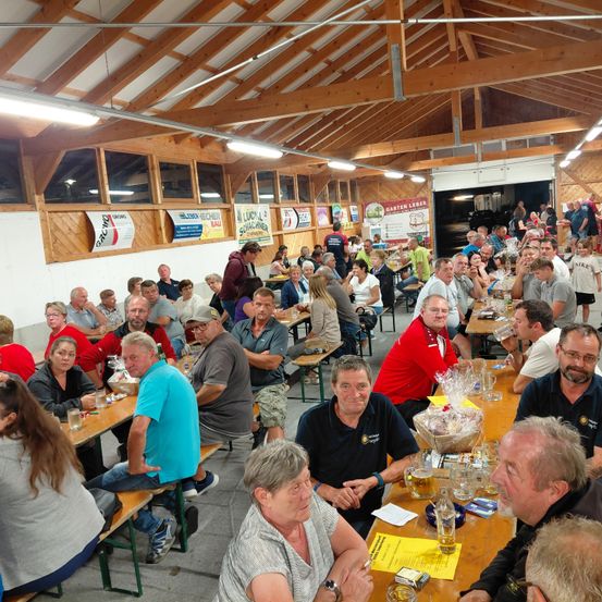 Eine große Gruppe von Menschen sitzt um Tische in einem Saal. Die Tische sind mit Gläsern, Tellern und verschiedenen Gegenständen gefüllt. Die Menschen sind lässig gekleidet. Der Saal hat Holzwände und Decken.
