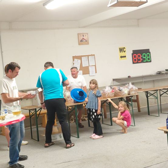 In einem Raum versammeln sich eine Gruppe Erwachsener und Kinder. Eine Anzeigetafel zeigt 89:98. Die Erwachsenen sind in einer Aktivität vertieft, während die Kinder zusehen. Auf den Tischen stehen Gläser und Flaschen.